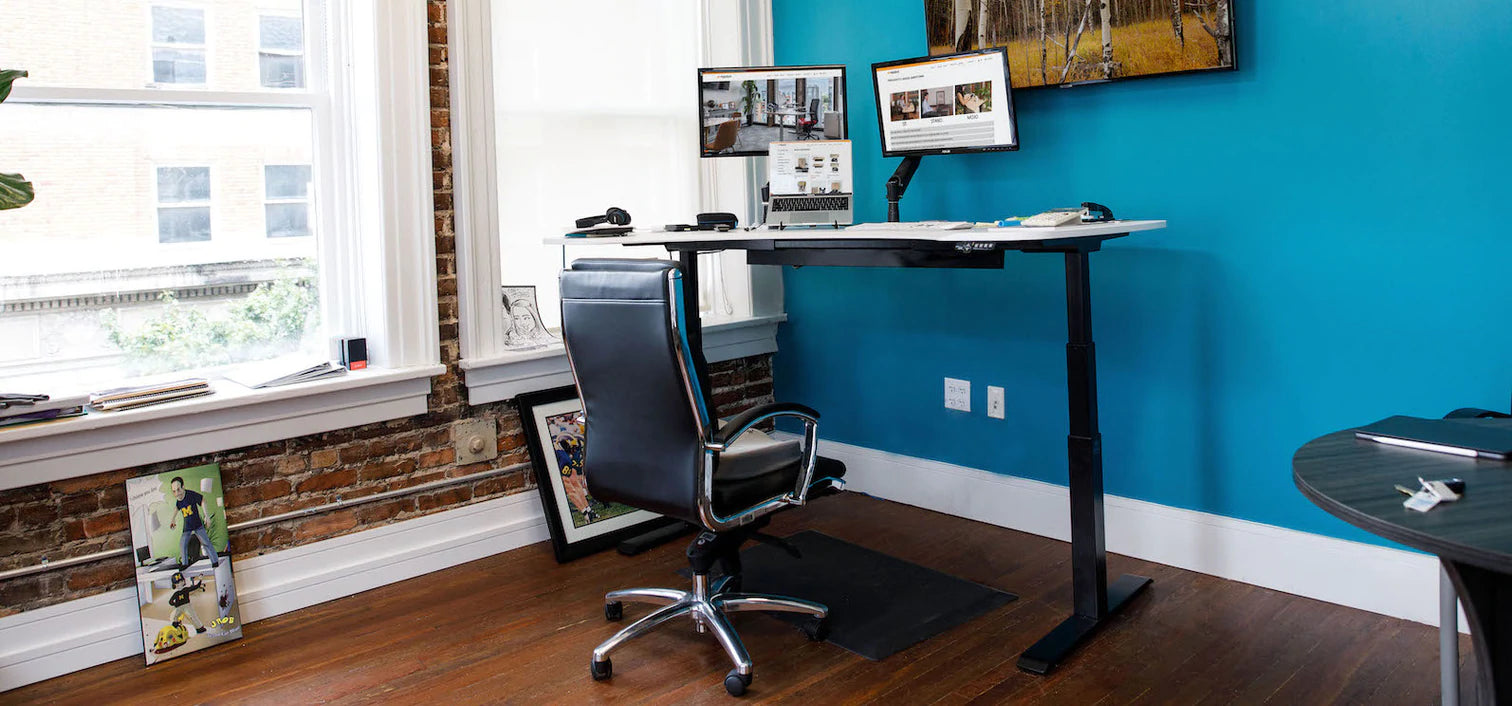 adjustable height stand up desk and black chair in front of blue wall with 2 monitor arms