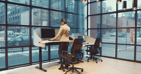 GIF showing an electric sit to stand desk with adjustable sound dampening panels adjust up and down with woman standing in front of it