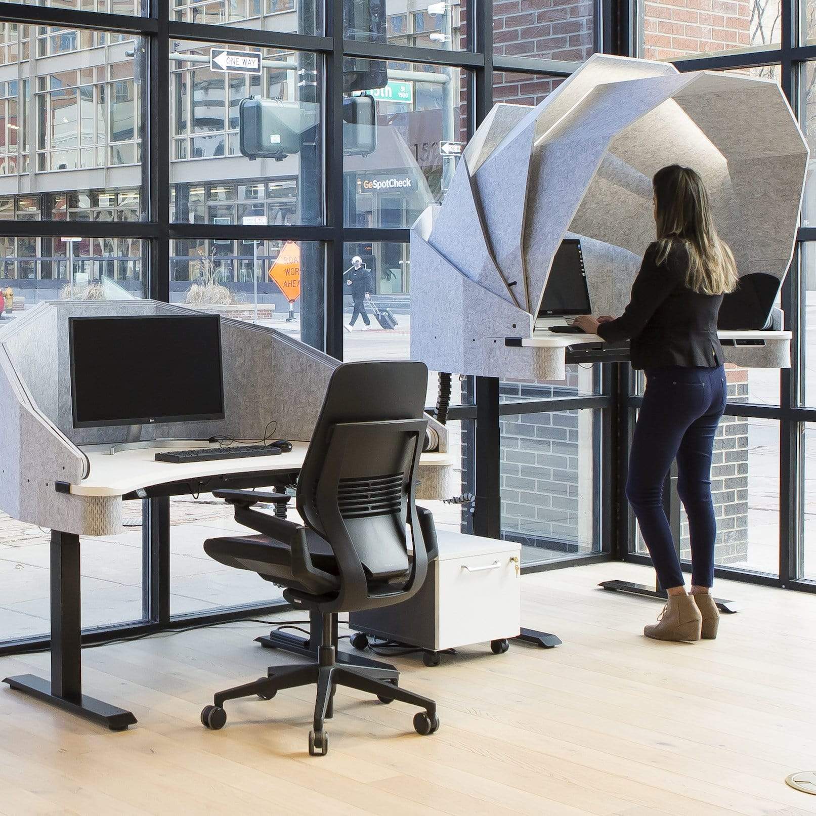2 MojoDome units shown side by side with electric standing desk sound absorbing panels up and down and woman standing typing on computer on the right hand side